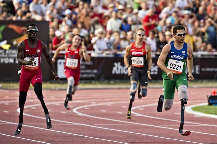 Em uma pista, quatro paratletas do atletismo paralímpico com próteses nas pernas estão correndo; na frente está um brasileiro, o Alan, com uma camisa meio azul meio verde, com o nome "Brasil"; ele está com uma bermuda verde e um óculos escuro de armação cinza.