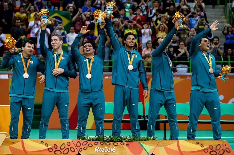 Os seis atletas brasileiros da seleção masculina de goalball estão no pódio da Rio-2016; estão sorridentes, com a medalha de bronze no pescoço e vestem um moletom verde escuro; ao fundo, o público aplaude.