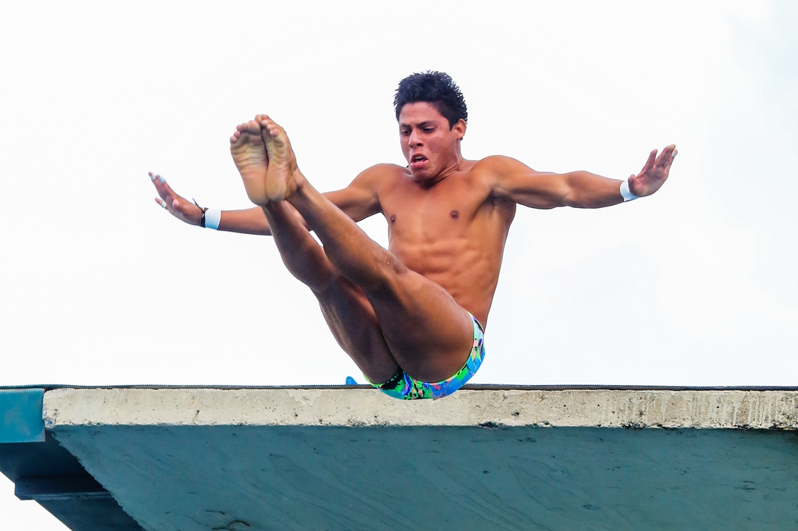 #PraCegoVer: o atleta Kawan Pereira compete em uma prova de saltos ornamentais. Na foto, ele está no ar após deixar a plataforma e em meio a um movimento com o corpo dobrado como se estivesse sentado, com os braços esticados para os lados em forma de cruz e as pernas estendidas para a frente